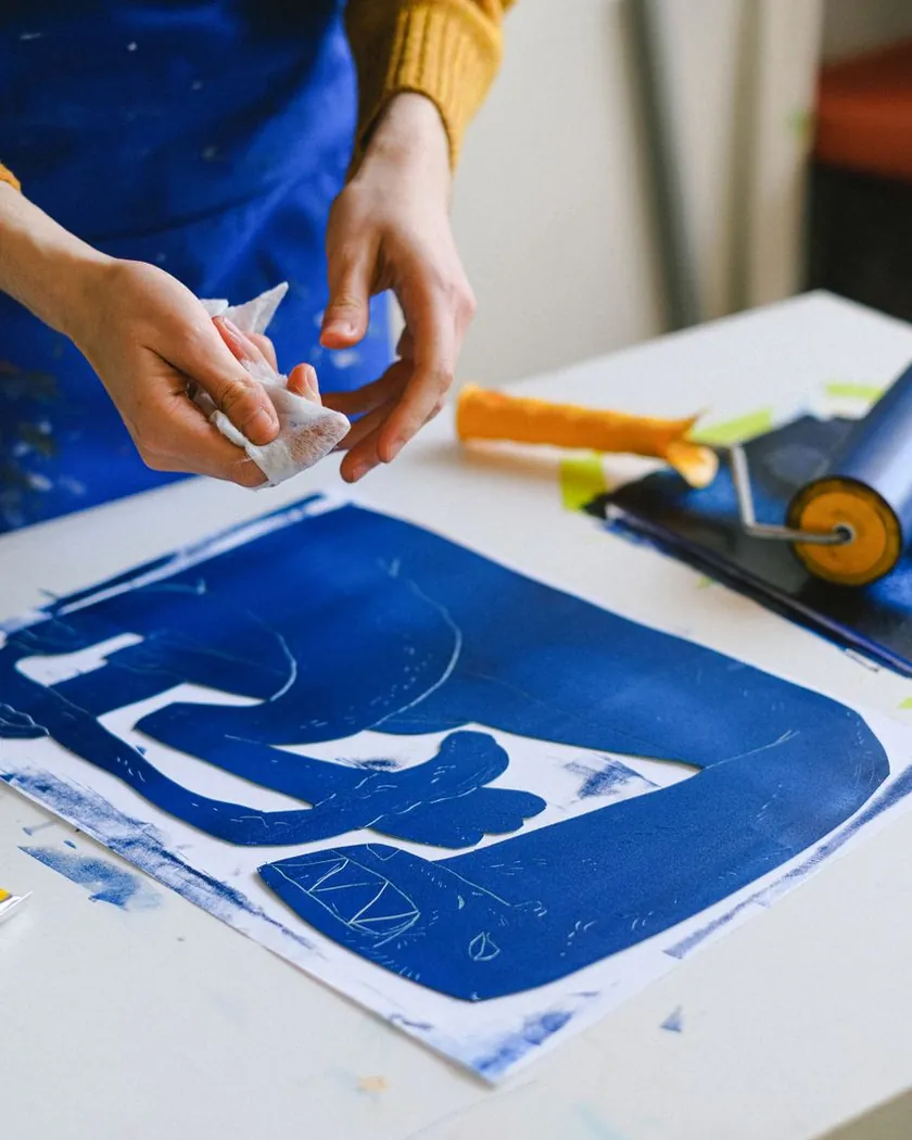 Manos limpiando con un paño una matriz de linóleo entintada en azul junto a un rodillo de estampación sobre la mesa del taller